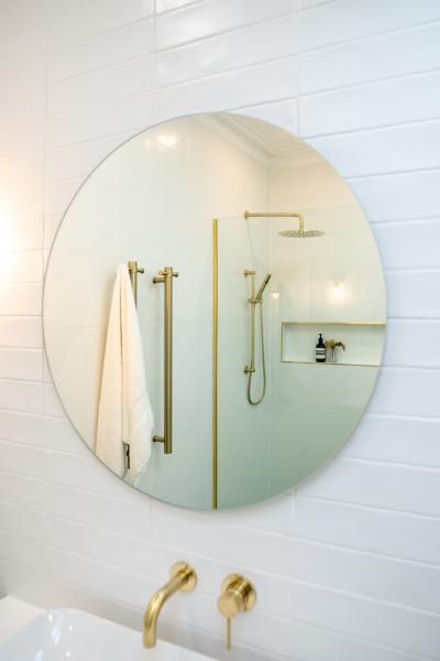 Round bathroom mirror above a white basin with a brass tap and shower reflected in the glass