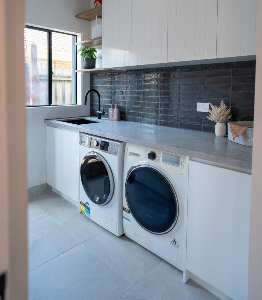 Modern laundry renovation with front-load washer, dryer, white cabinetry and dark tiled splashback