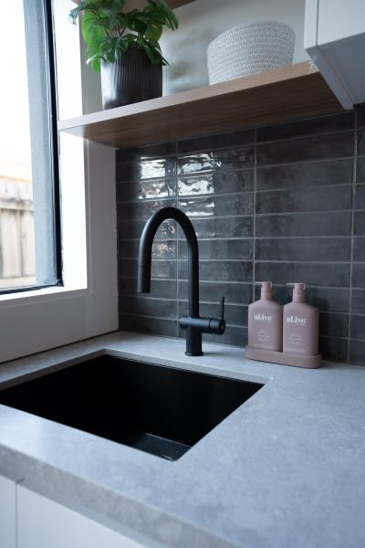 Matte black kitchen tap and sink with grey tiled splashback beside a window