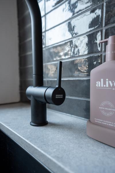 Matte black bathroom tap beside pink soap bottle on grey vanity with glossy tiled splashback