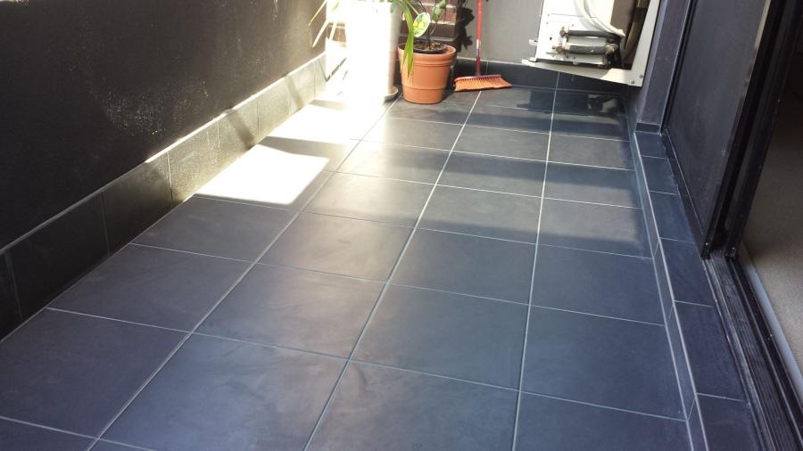 Dark floor tiles installed along a narrow balcony walkway with sunlight and sliding doors
