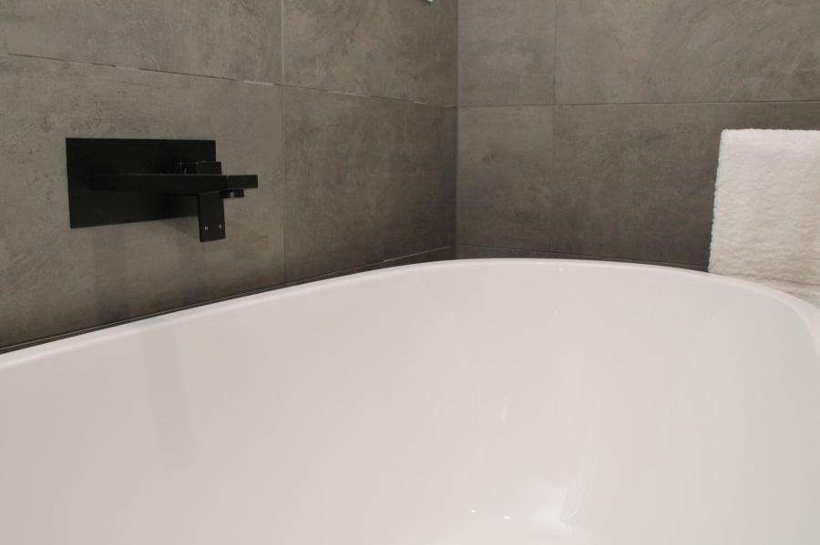Close-up of a white freestanding bath with matte black wall tap against grey tiled bathroom wall