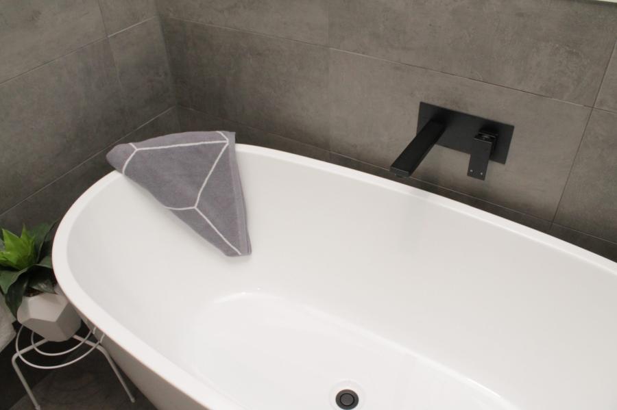 Freestanding white bath with black wall-mounted tapware against grey tiled bathroom walls