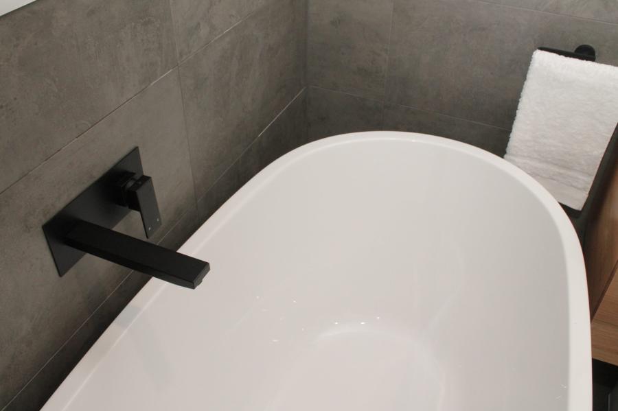 Freestanding white bath with matte black wall-mounted bath spout against grey tiled wall