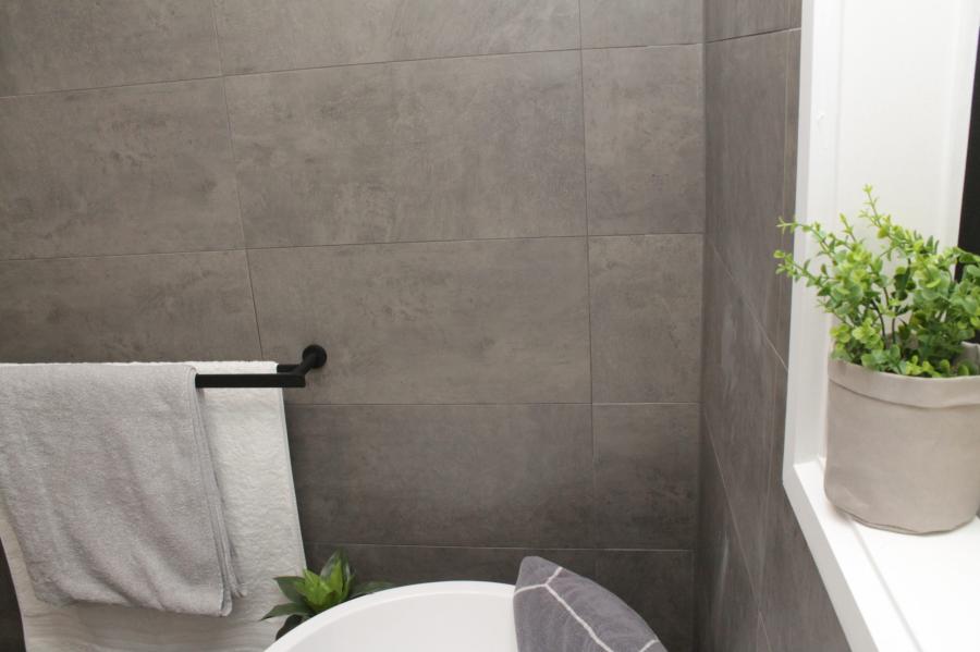 Grey tiled bathroom corner with black towel rail, white towel, plant and bath edge