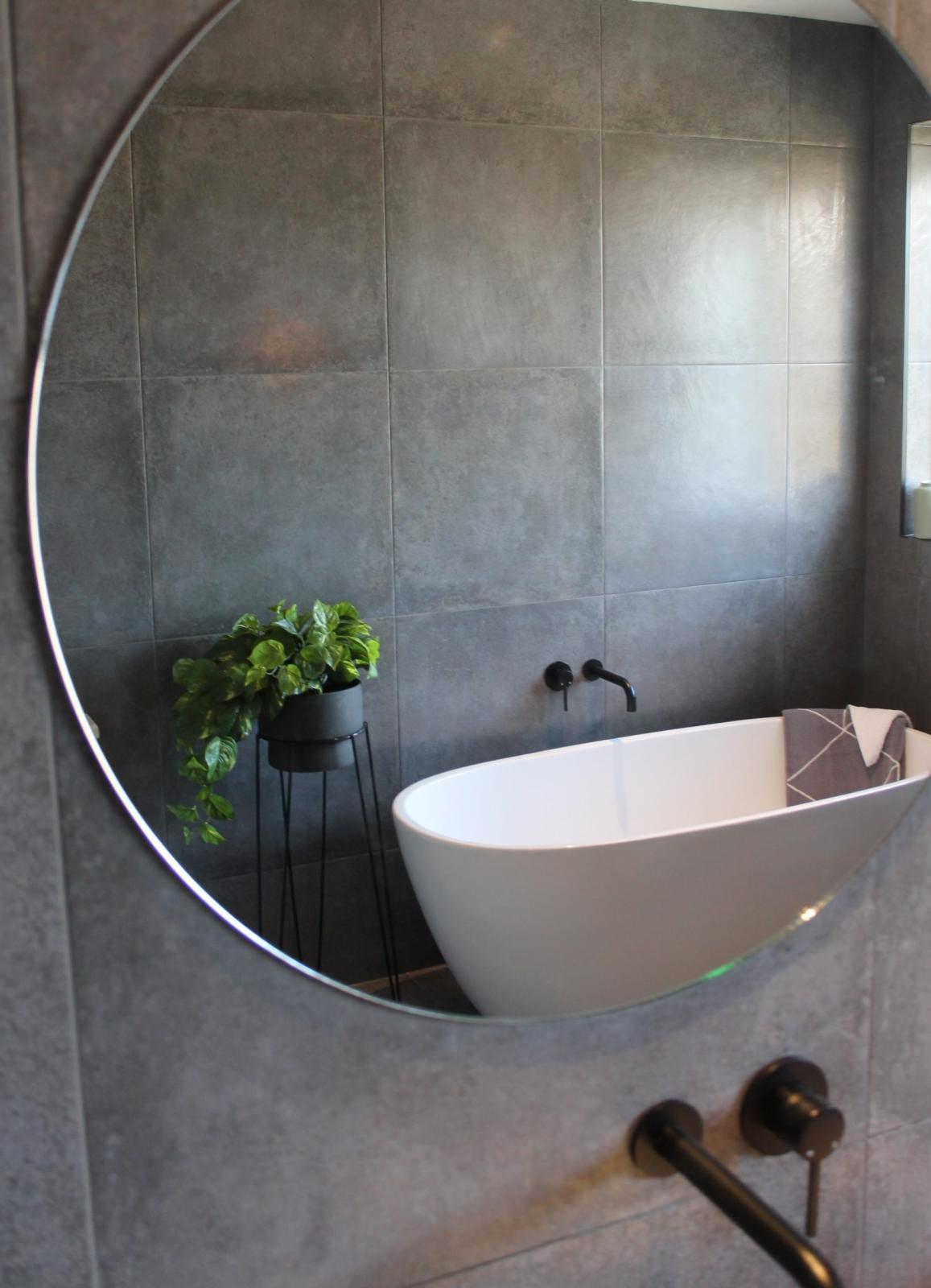 Freestanding white bath reflected in a round mirror against grey tiled bathroom walls