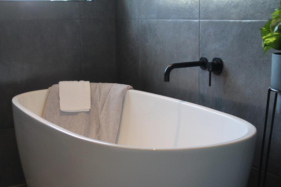 Freestanding white bath with black wall tapware against dark grey tiled bathroom wall