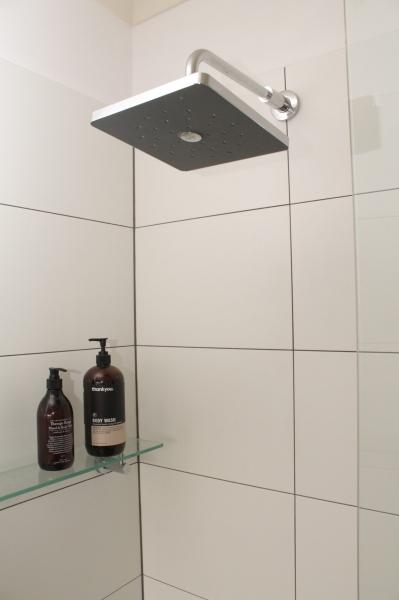 Square rainfall shower head above a tiled shower wall with glass shelf and toiletries