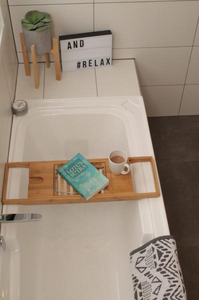 White bathtub with timber bath caddy, book, mug and plant in a modern tiled bathroom