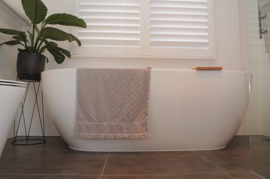 Freestanding white bathtub with folded towel, indoor plant and shuttered window in a modern bathroom
