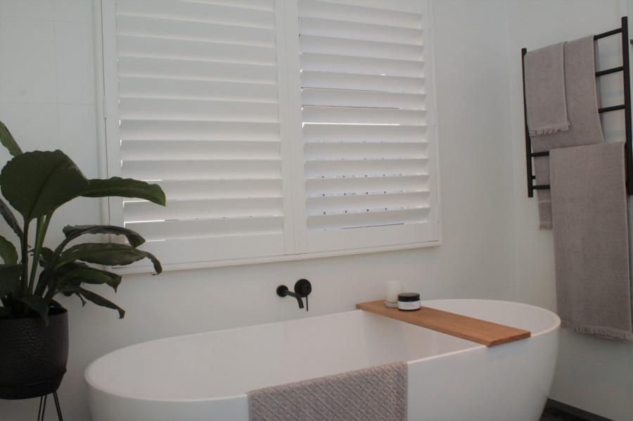 Freestanding white bathtub beneath shuttered window with timber bath caddy and black towel rail