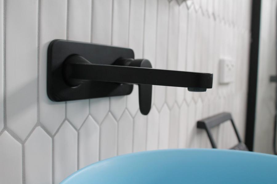 Matte black wall-mounted bathroom tap on white feature tiles above a blue basin