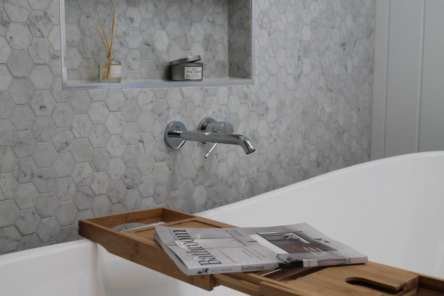 Freestanding bath with timber bath caddy beside white hexagon tiled bathroom wall niche