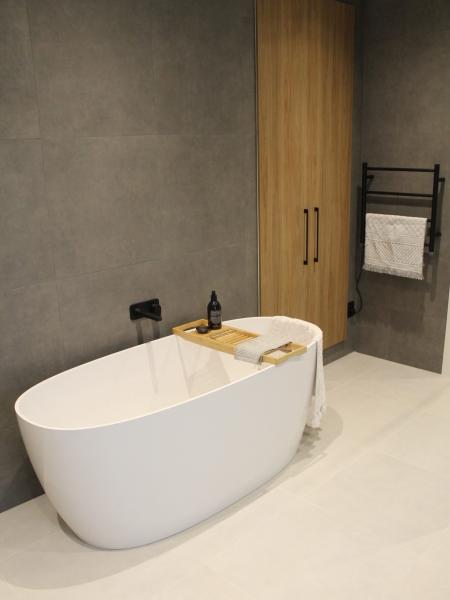 Modern freestanding white bath with grey tiled wall and timber cabinetry