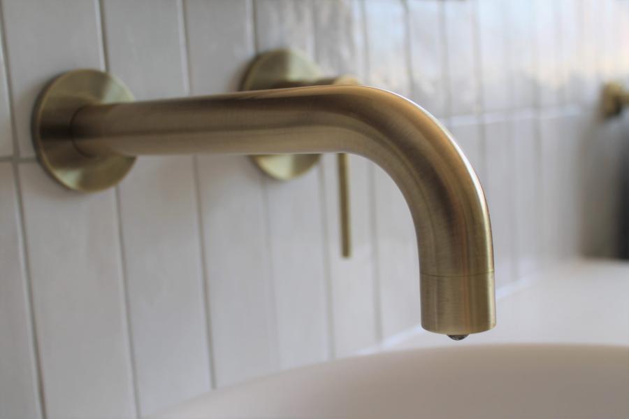 Brushed brass wall-mounted bathroom tap above a white basin and tiled splashback