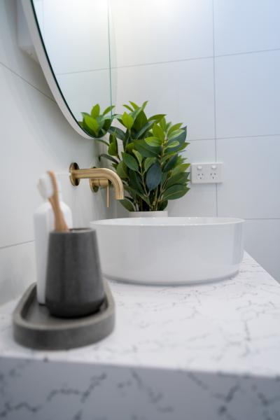 Brushed brass wall tap above a white basin on a marble-look vanity with mirror and greenery