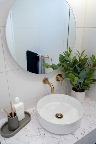 Round bathroom mirror above white basin with brass tapware and potted greenery