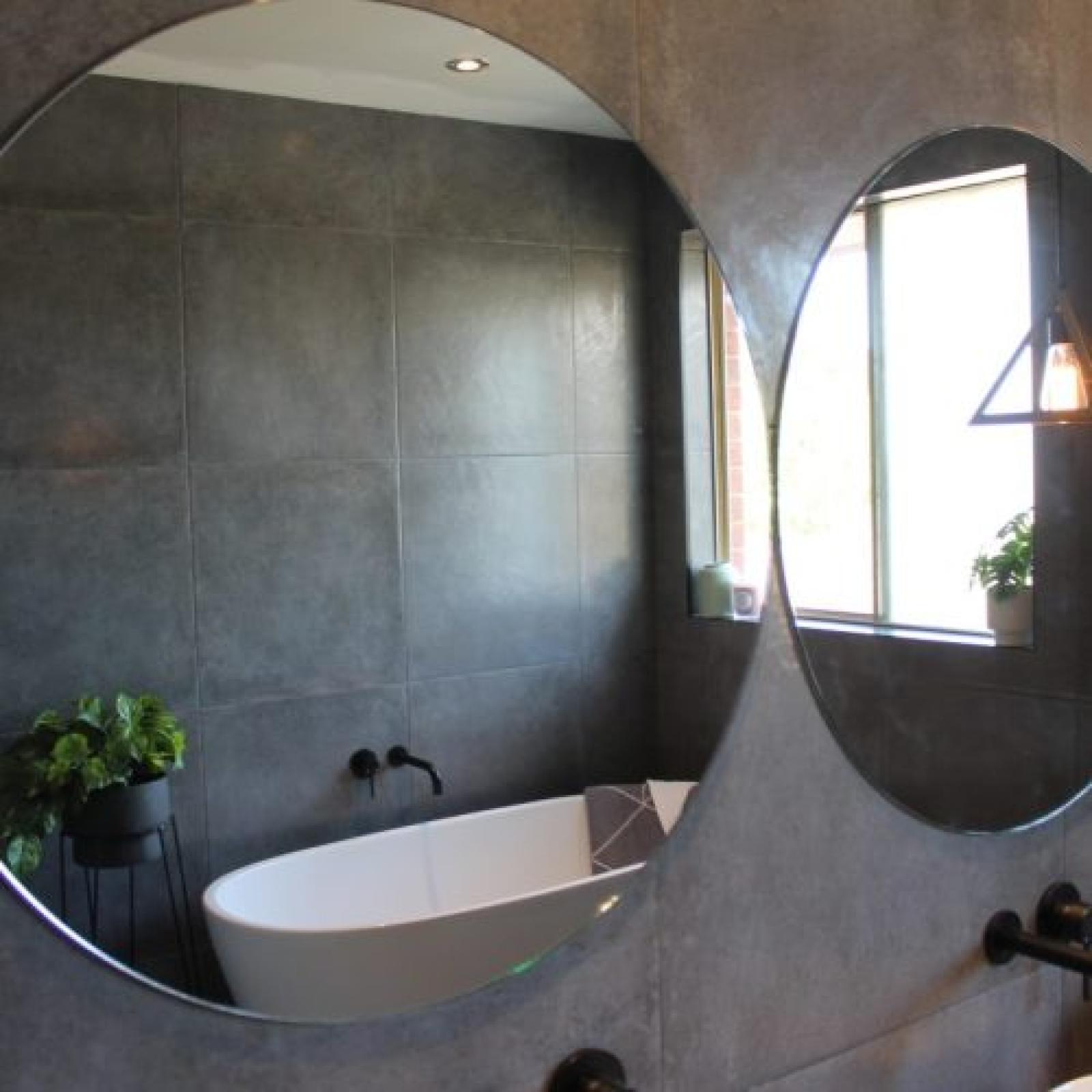 Round mirror reflecting a modern bathroom with freestanding bath and grey tiled walls